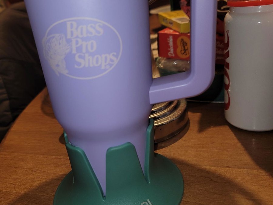 A lavender Bass Pro Shops insulated mug with a handle seated in a green drink base marked STEADI, standing on a cluttered wooden side table with jars and a red cup behind